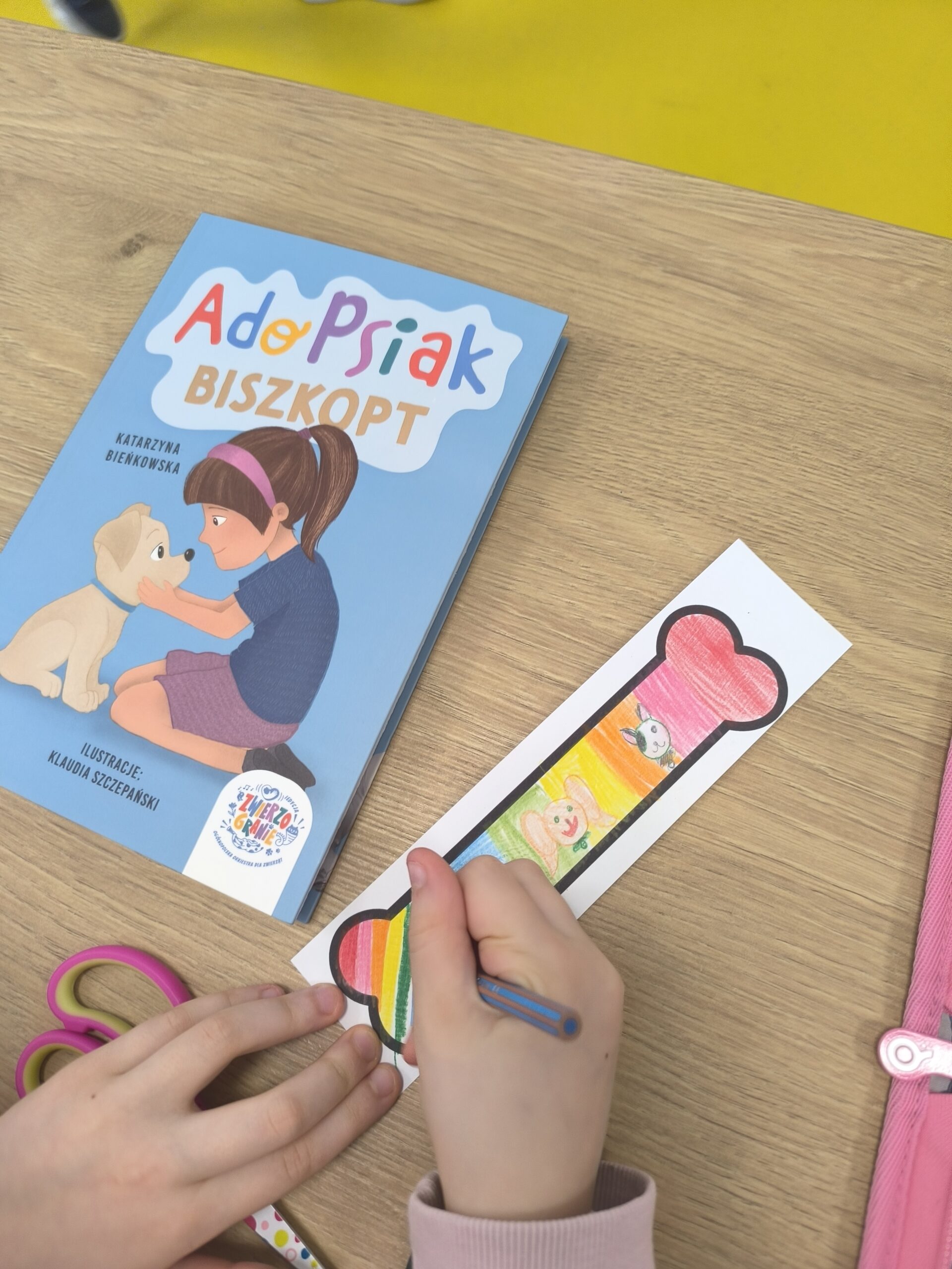 Dziecko koloruje zakładkę w kształcie kości obok książki AdoPsiak Biszkopt
